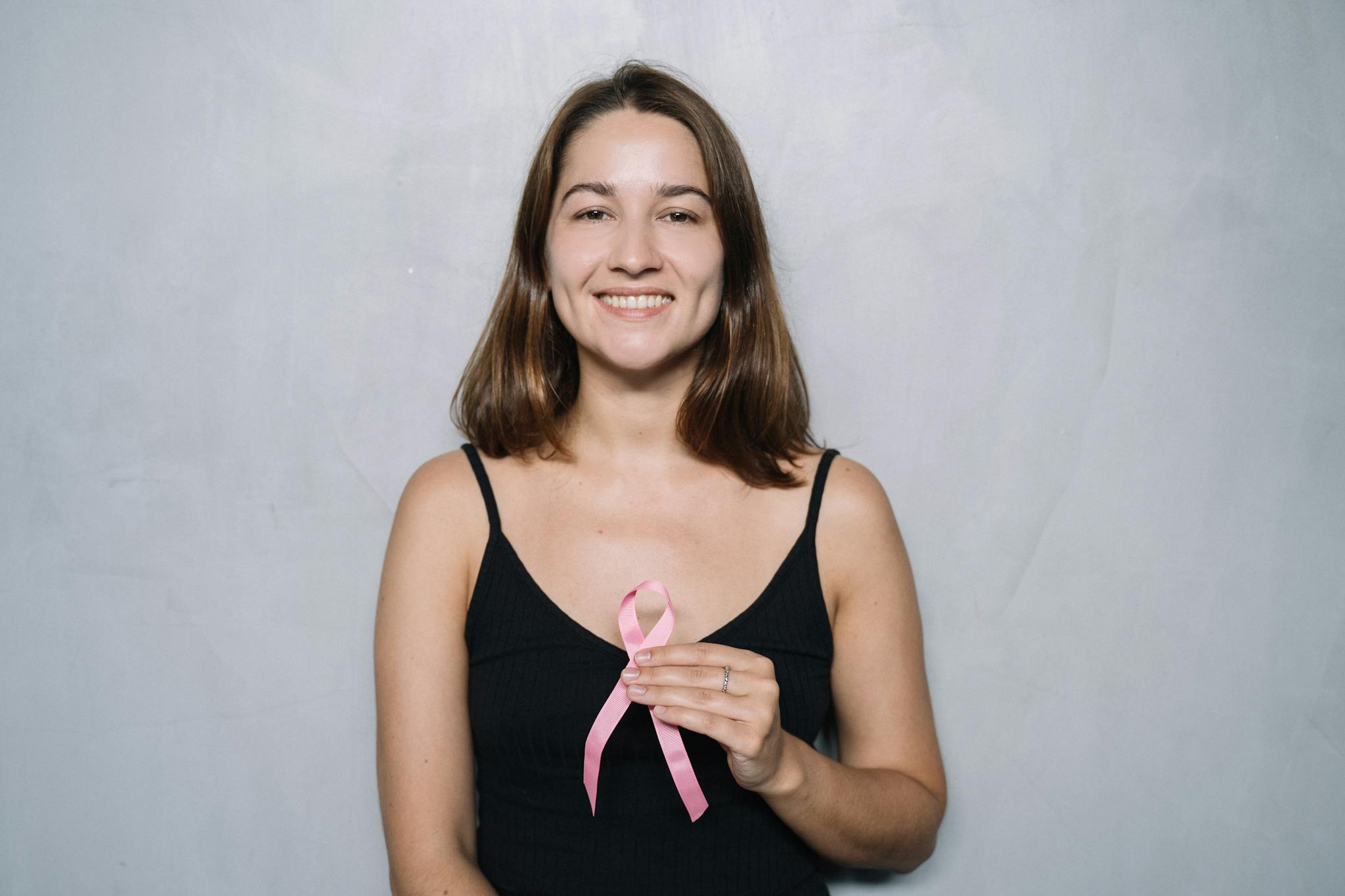 Smiling woman holding a pink ribbon symbolizing breast cancer awareness against a neutral background.